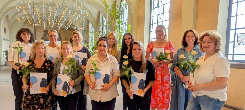 Frauen stehen Zertifikaten und Rosen ind en Händen in zwei Reihen in einem Raum.