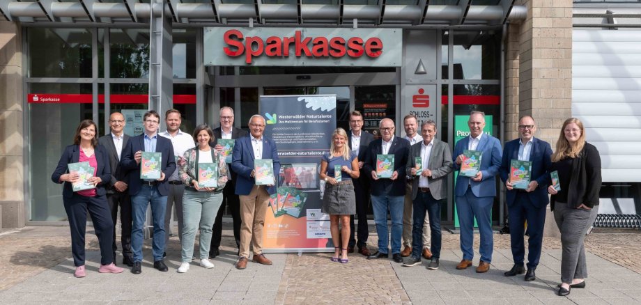 Frauen und Männer stehen vor der Sparkasse und halten jeweils eine Ausbildungsfibel in der Hand.
