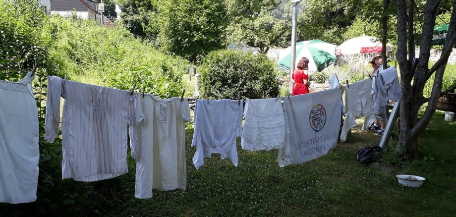 Auf einer Wäscheleine im Garten hängen große, historisch anmutende weiße Kleiderstücke.