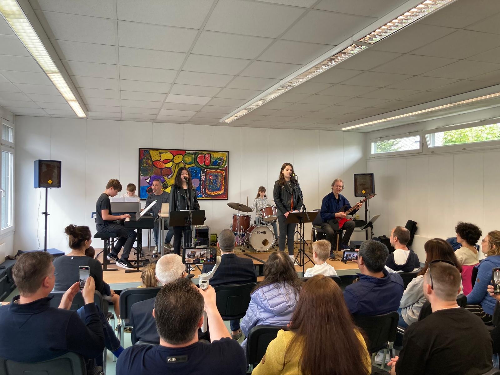 Auf einer Bühne musizieren Schülerinnen und Schüler mit verschiedenen Instrumenten vor einem Publikum.