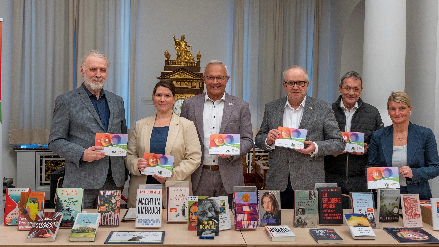 Vier Männer und zwei Frauen stehen mit Programmheften in der Hand hinter einem Büchertisch.
