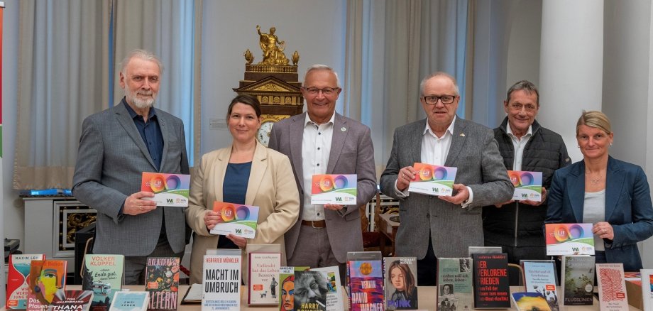 Vier Männer und zwei Frauen stehen mit Programmheften in der Hand hinter einem Büchertisch.