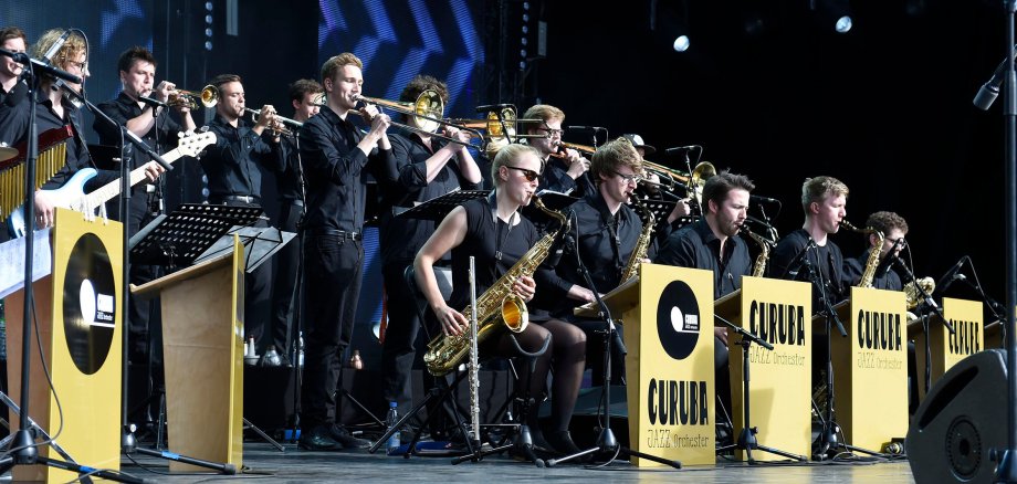 Auf einer Bühne stehen und sitzen Musizierende des Jazz-Orchesters Curuba mit ihren Instrumenten.