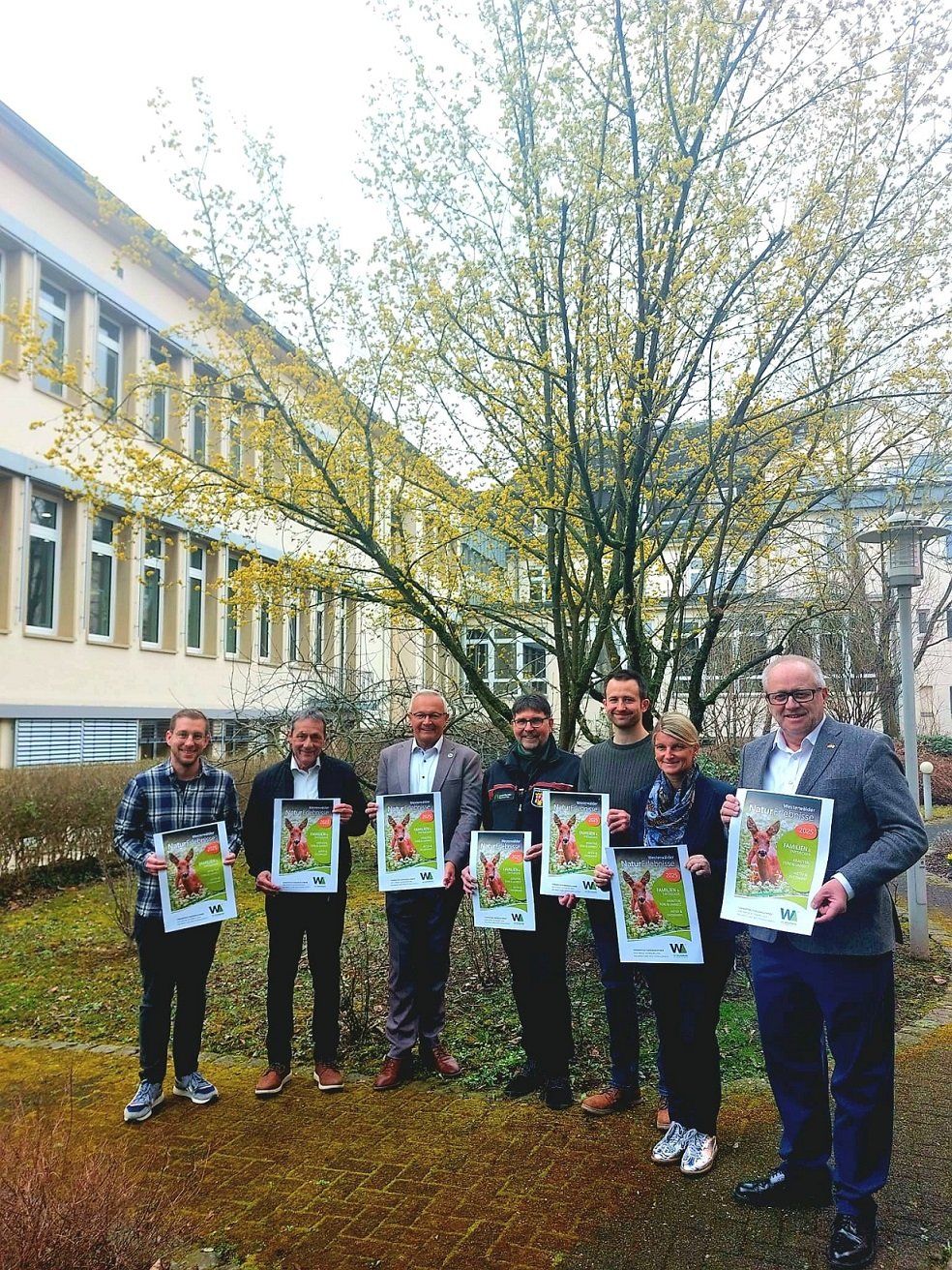 Sechs Männer und eine Frau stehen mit einem Plakat der Naturerlebnisse vor einem Baum im Innenhof.