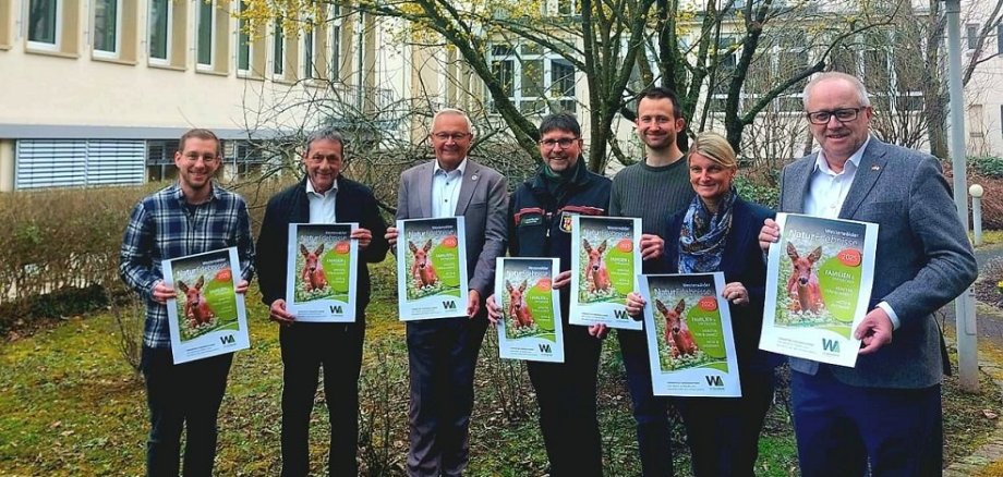 Sechs Männer und eine Frau stehen mit einem Plakat der Naturerlebnisse vor einem Baum im Innenhof.