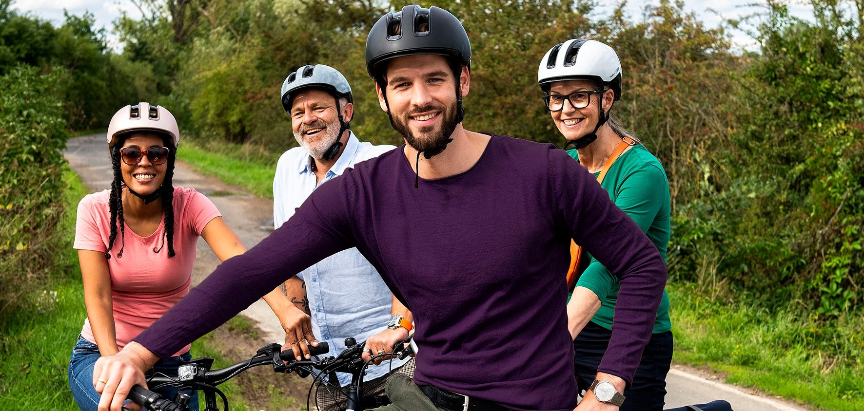 Vier Fahrradfahrer, zwei Männer und zwei Frauen, mit Fahrradhelm stehen im Grünen und lachen in die Kamera.