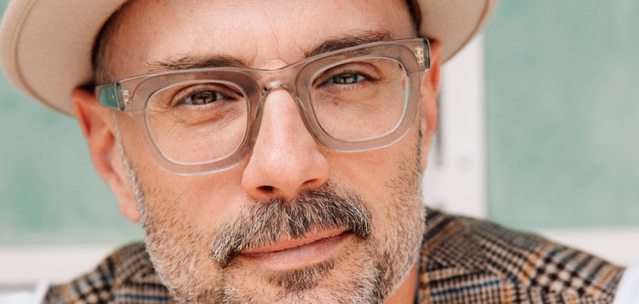 In dem Porträt ist ein bärtiger Mann mit Brille und Hut sowie karierter Weste zu sehen.