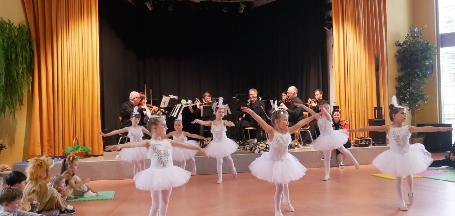 Mädchen in weißen Ballettkleidchen tanzen als Schwäne, im Hintergrund ist ein Orchester.