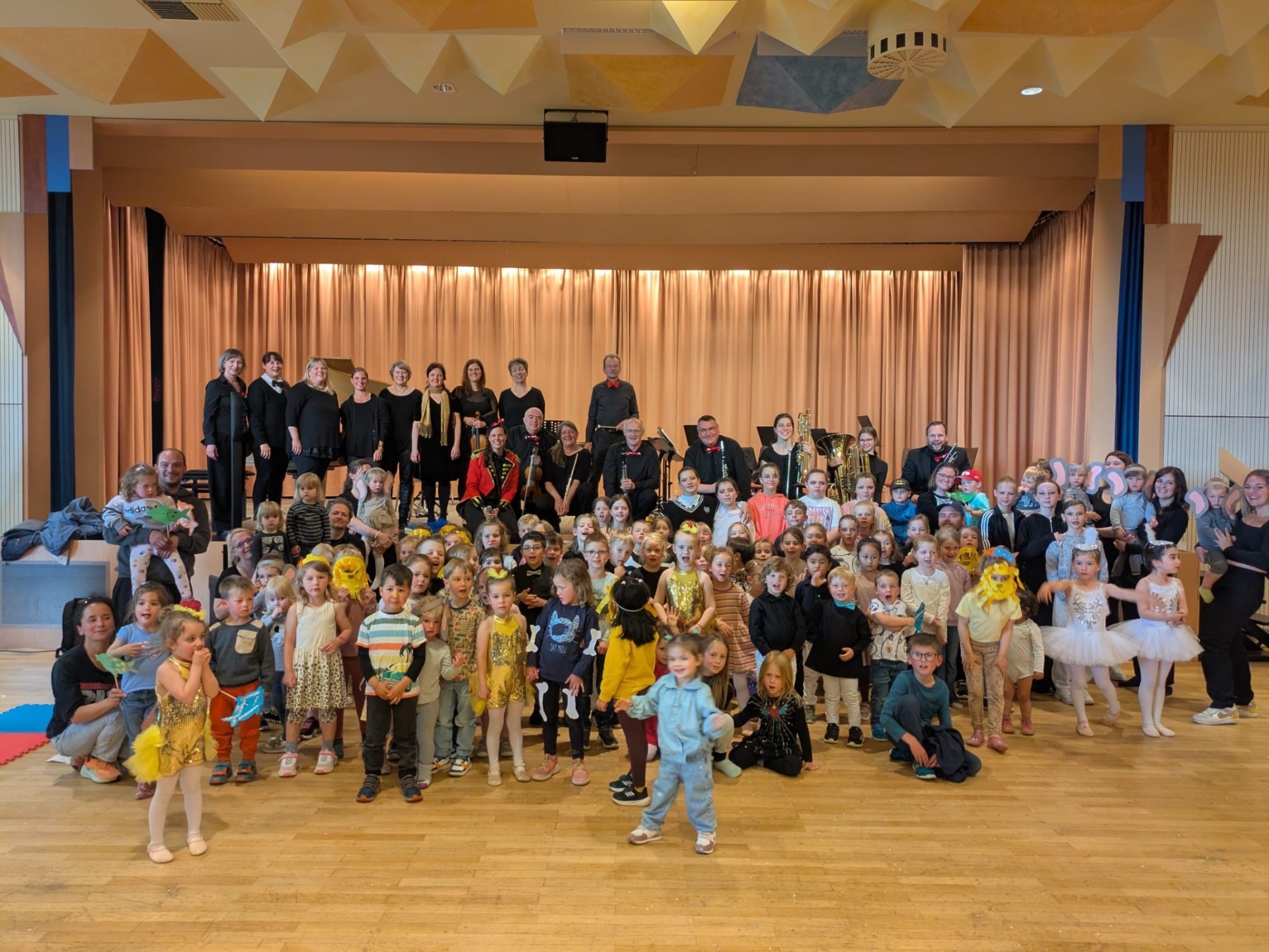 Eine bunt kostümierte Kinderschar steht vor einer Bühne, auf der ein Orchester steht.