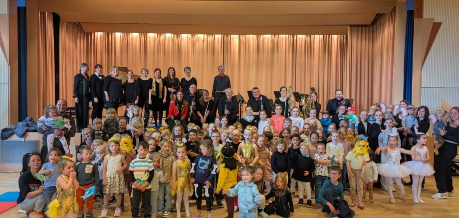 Eine bunt kostümierte Kinderschar steht vor einer Bühne, auf der ein Orchester steht.