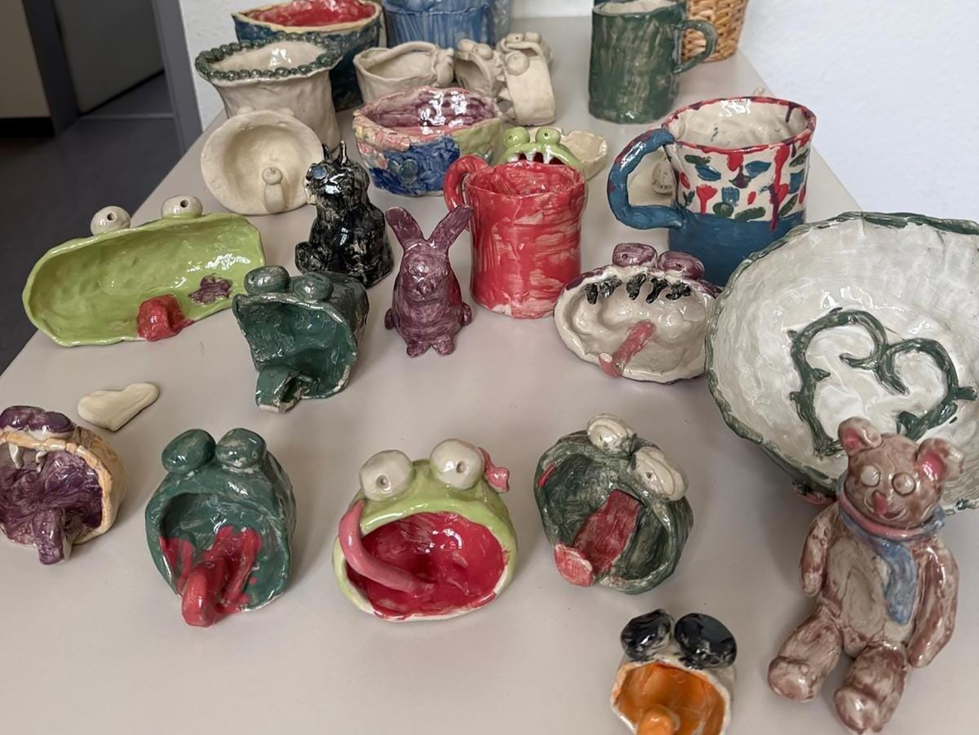 Auf einem Tisch stehen bunte Keramikwerke, darunter Frösche mit offenem Maul und langer Zunge, Hasen, Teddys, Vasen und Becher.