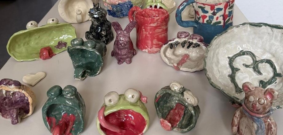 Auf einem Tisch stehen bunte Keramikwerke, darunter Frösche mit offenem Maul und langer Zunge, Hasen, Teddys, Vasen und Becher.