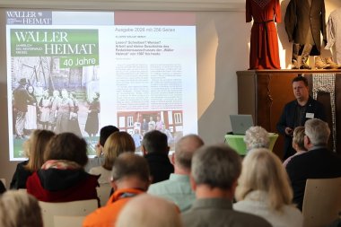 Auf einer Leinwand ist die aktuelle Ausgabe der Wäller Heimat zu sehen. Rechts davon steht ein mann am Rednerpult und spricht zu den sitzenden Personen vor ihm.