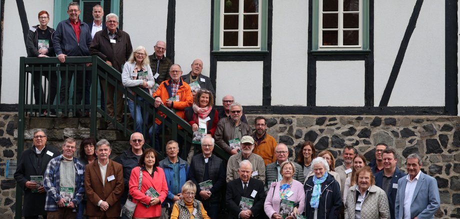 Vor einem Fachwerkhaus mit Treppe steht eine große Gruppe Menschen.