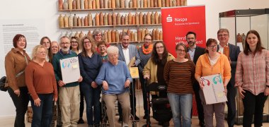 Beschäftigte der Caritas Werkstätten, Mitarbeitende des Museums und die offiziellen Ehrengäste stehen vor einer Wand mit zahlreichen Tonkrügen.
