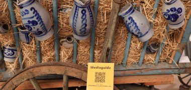 Vor einem alten Leiterwagen mit Kannen daran steht ein gelbes Schild mit Mediaguide und dem Link https://keramikmuseum.pwa.tonwelt.com darauf.