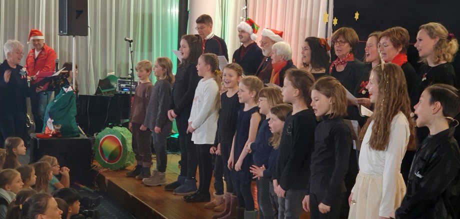 Auf einer Bühne stehen Kinder und Erwachsene, zum Teil im weihnachtlichen Outfit, und singen.