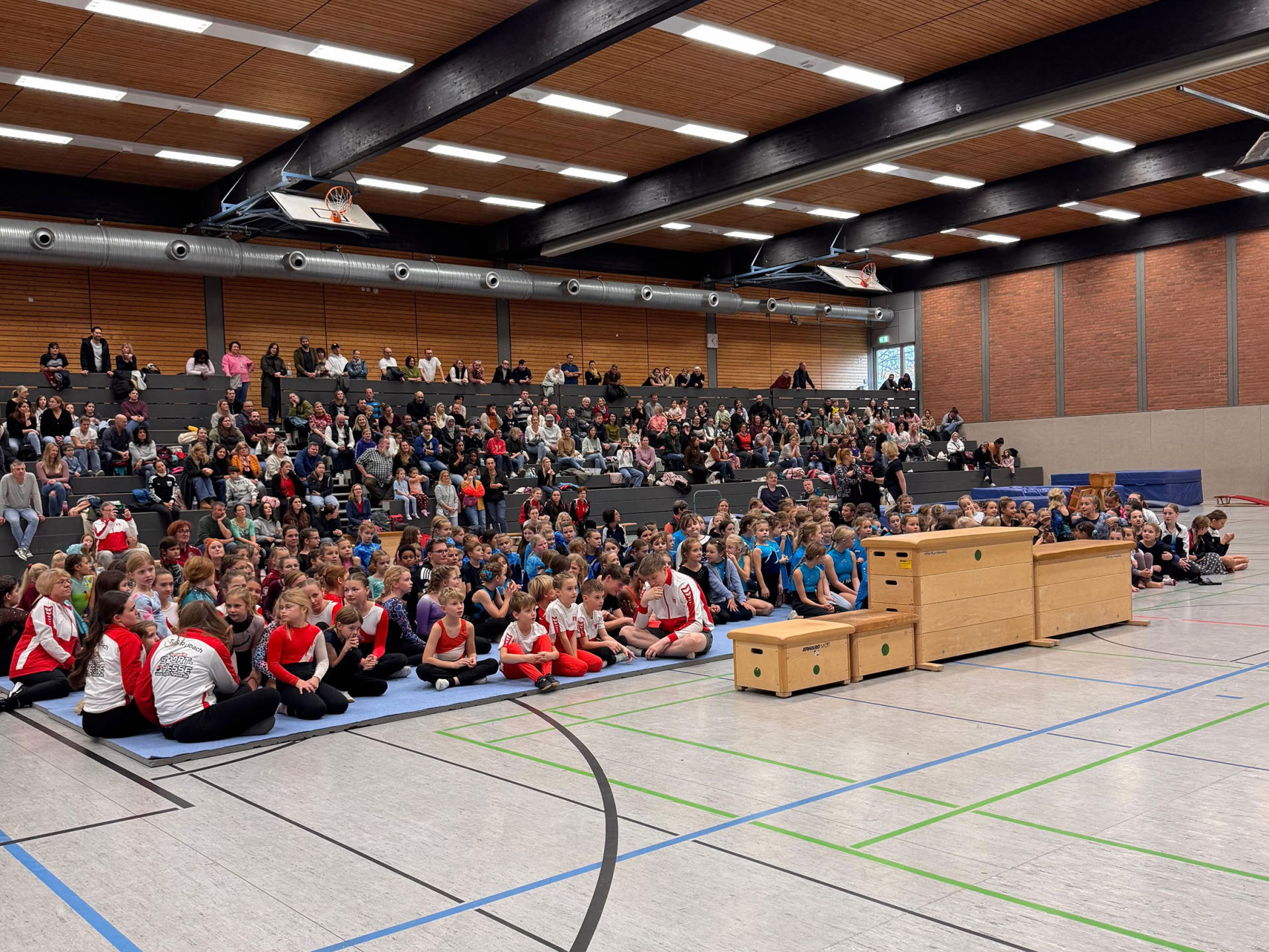 In einer Turnhalle sitzen vorn auf Matten zahlreiche Kinder in Sportkleidung und dahinter auf Rängen Erwachsene.