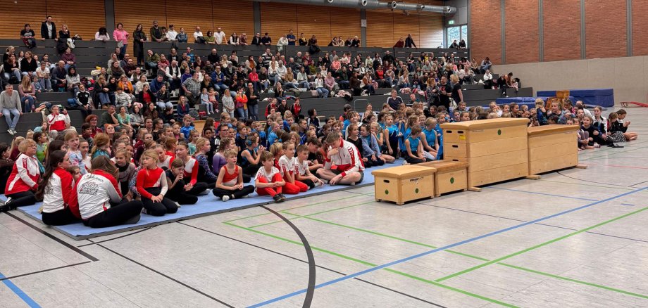 In einer Turnhalle sitzen vorn auf Matten zahlreiche Kinder in Sportkleidung und dahinter auf Rängen Erwachsene.
