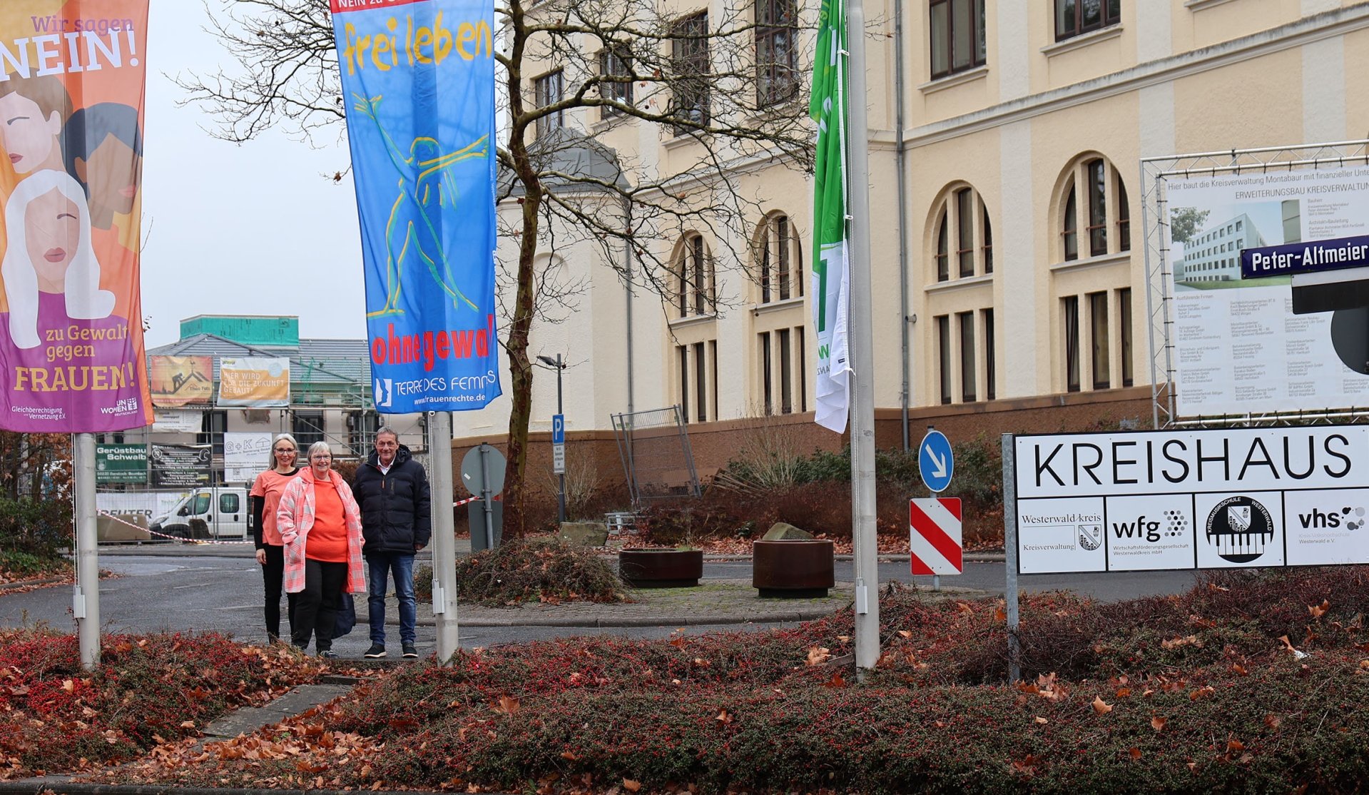Vor dem Gebäude der Kreisverwaltung stehen zwei in orange gekleidete Frauen und ein Mann zwischen zwei gehissten Fahnen mit der Aufschrift "Wir sagen nein! zu Gewalt gegen Frauen" und "frei leben ohne Gewalt". 