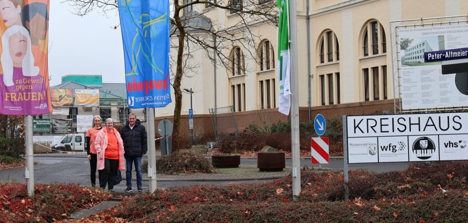Vor dem Gebäude der Kreisverwaltung stehen zwei in orange gekleidete Frauen und ein Mann zwischen zwei gehissten Fahnen mit der Aufschrift "Wir sagen nein! zu Gewalt gegen Frauen" und "frei leben ohne Gewalt".