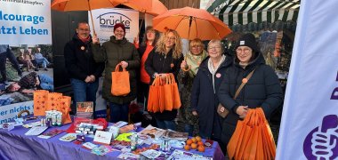 Mehrere Frauen und ein Mann stehen mit orangenen Regenschirmen und Stoffbeuteln an einem Infostand auf einem Marktplatz.