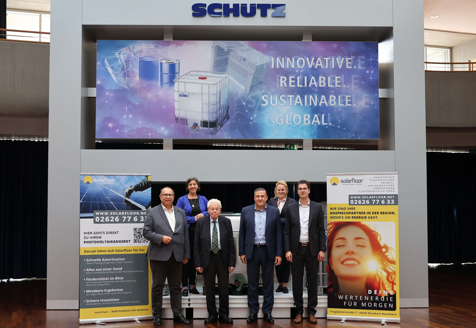Zwei Frauen und vier Männer in Anzügen stehen vor einer Werbewand der Firma Schütz mit Roll-ups von solarfloor.