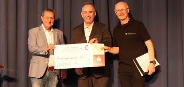 Auf der Bühne erfolgt die Scheckübergabe der drei Männer.