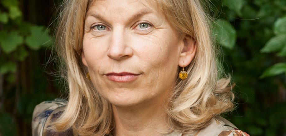 Eine Frau mit schulterlangem blonden Haar und einer geblümten Bluse sitzt im Grünen.
