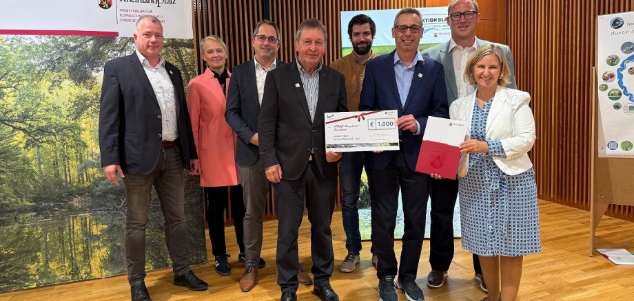Zwei Frauen und sechs Männer stehen vor einem Ministeriumsbanner, zwei Männer halten einen Scheck über 1.000 Euro in den Händen.