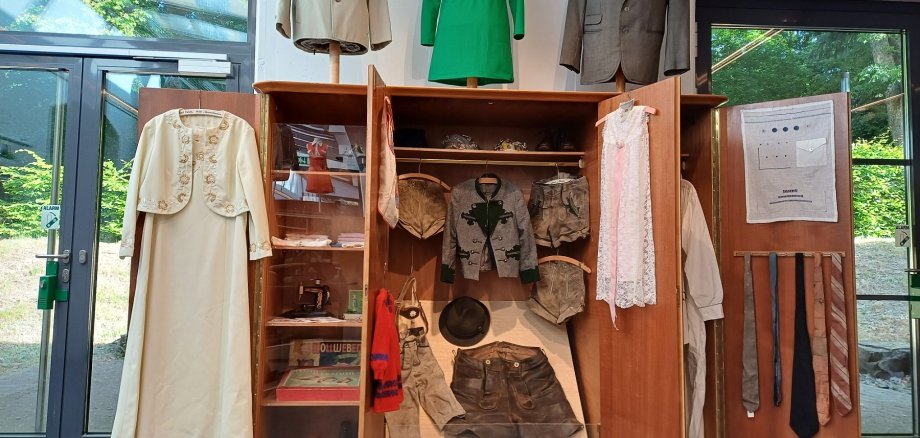 In einem alten geöffneten Kleiderschrank hängen mehrere alte Kleidungsstücke, darunter Kleider, Jacketts und Blusen.