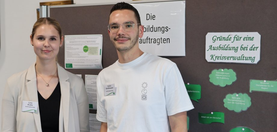 Eine junge Frau und ein junger Mann stehen lächelnd vor einer Infowand.