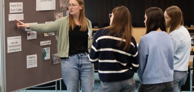 Eine langhaarige junge Frau erklärt drei Mädchen  Informationen zum dualen Studiengang "Soziale Arbeit" an einer Infowand.