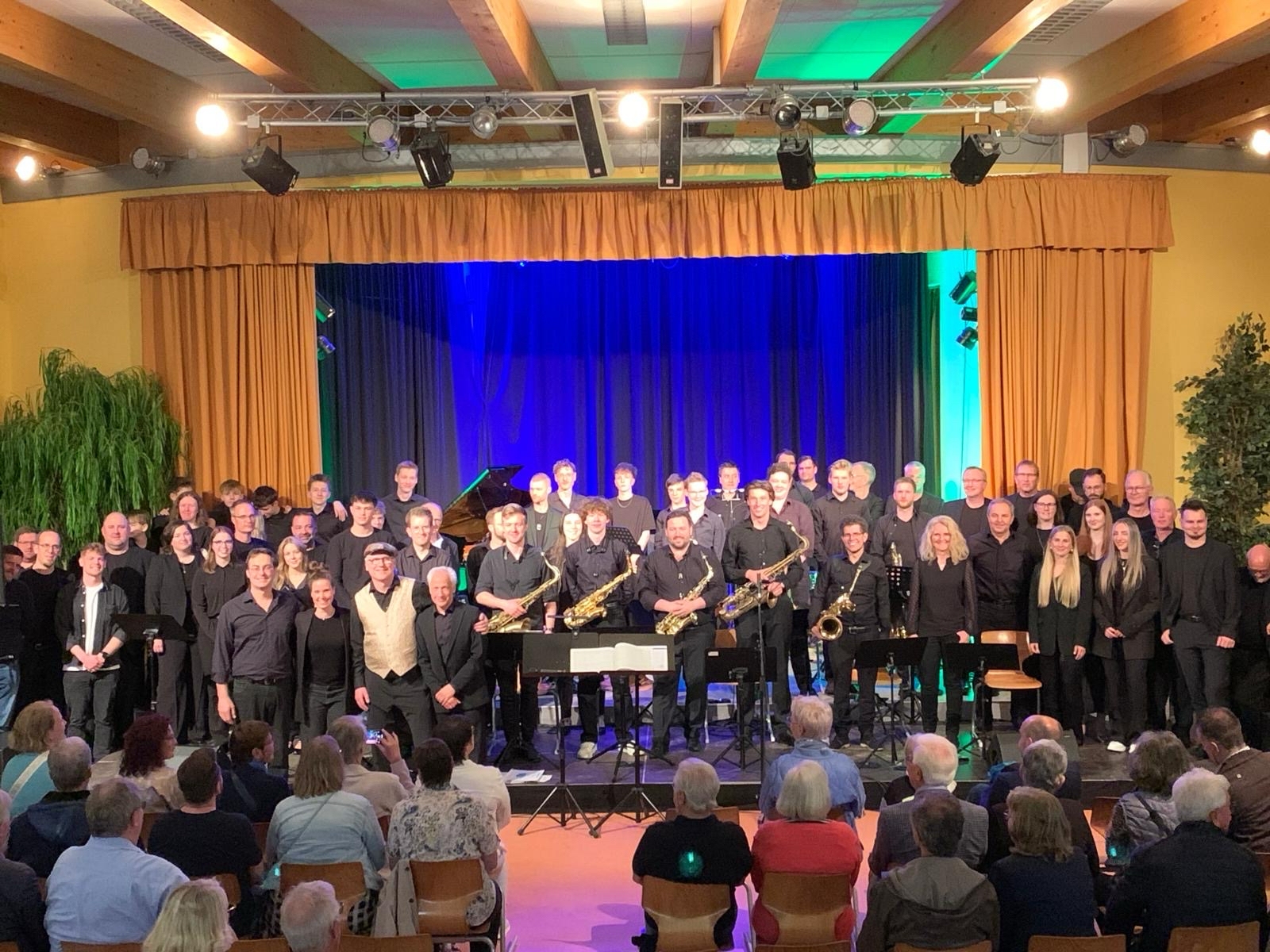 Eine große Gruppe schwarzgekleideter Personen steht, teilweise mit Instrumenten, vor Publikum auf einer Bühne.