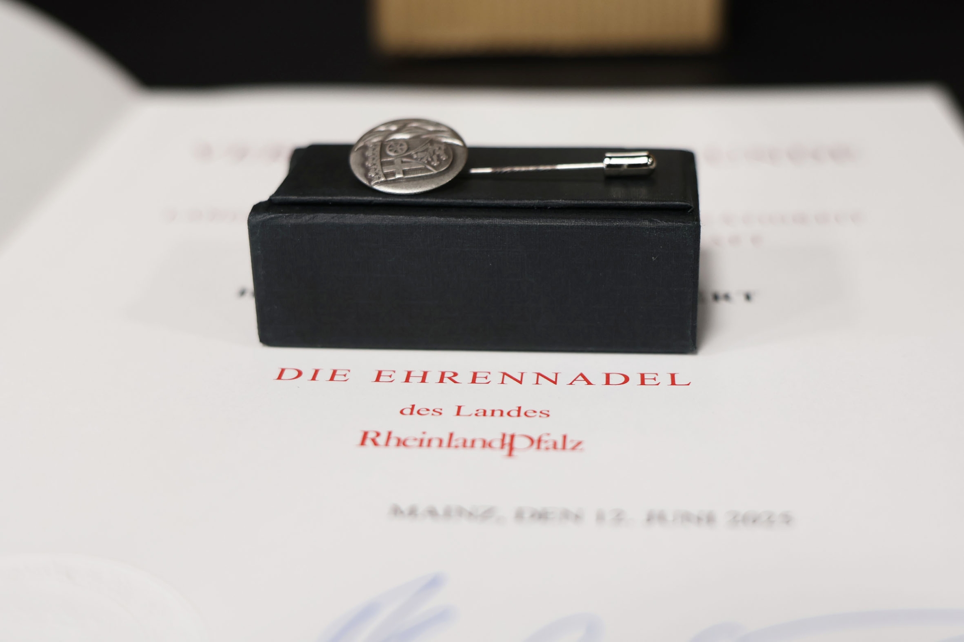 Die Ehrennadel liegt auf einem schwarzen Kästchen, darunter die Urkunde des Landes Rheinland-Pfalz.