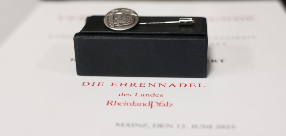 Die Ehrennadel liegt auf einem schwarzen Kästchen, darunter die Urkunde des Landes Rheinland-Pfalz.