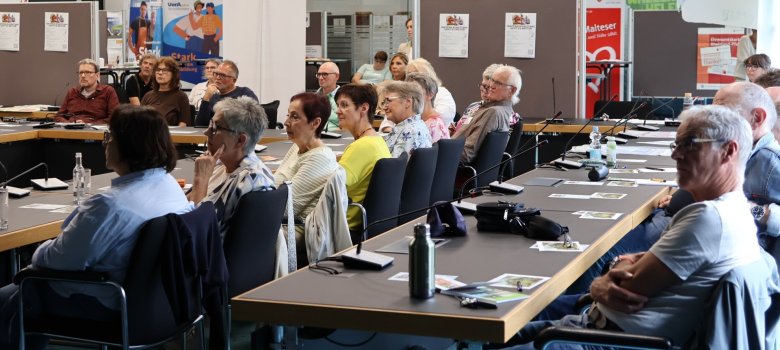 Im Veranstaltungsraum sitzen intterssierte Seniorinnen und Senioren an Tischen. Im Hintergrund ist eine Ausstellung zu sehen.