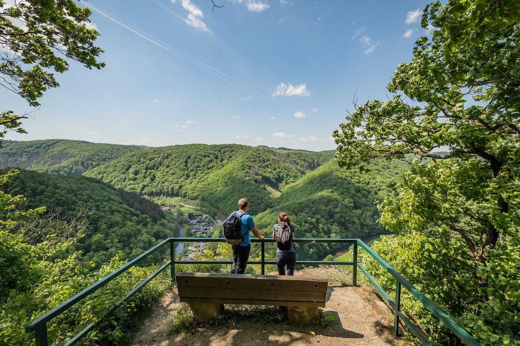 Zwei Wanderer stehen auf einer Aussichtsplattform mit Weitblick über ein grünes Tal mit Fluss.