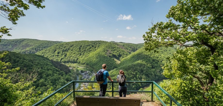 Zwei Wanderer stehen auf einer Aussichtsplattform mit Weitblick über ein grünes Tal mit Fluss.