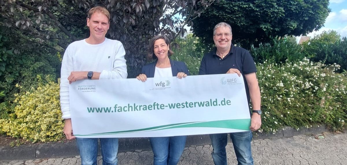 Zwei Männer und eine Frau stehen im Grünen mit einem Banner, auf dem www.fachkraefte-westerwald.de steht.