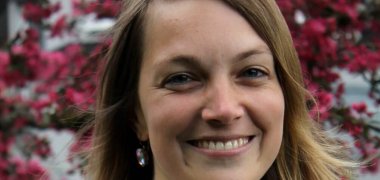 Eine junge Frau steht lächelnd vor einem rot blühenden Baum.