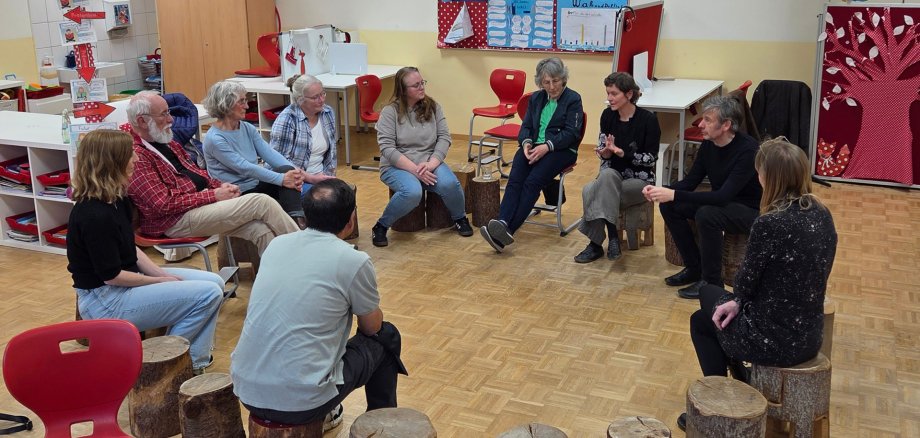 Frauen und Männer sitzen auf Baumstammhockern in einem Klassenraum.