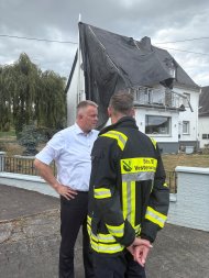 Der Ministerpräsident steht mit einem Feuerwehrmann vor einem Haus ohne Dach.