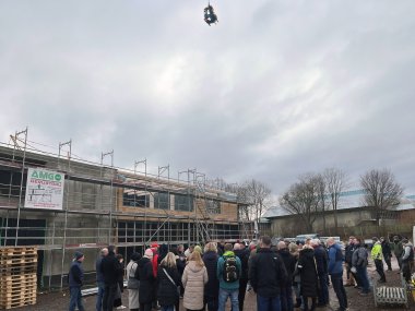 Über einem Rohbau und Menschen davor schwebt eine Krone aus Tannenzweigen.