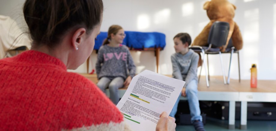 Eine Frau mit Textblatt übt mit zwei Kindern etwas ein.