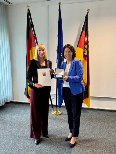 Zwei Frauen stehen mit Urkunde und Medaille vor drei Fahnen.