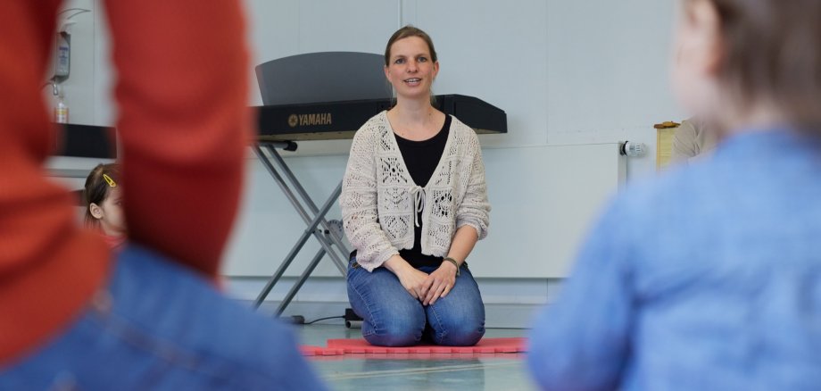 Eine Frau kniet auf einer Moosgummimatte und singt vor Kindern.