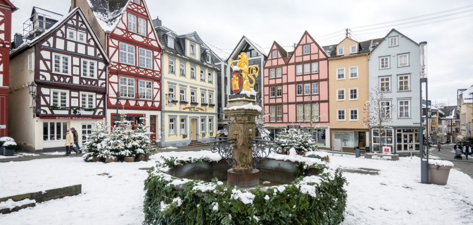 Ein mit Tannenzweigen geschmückter Brunnen im Winter vor Fachwerkhäusern.