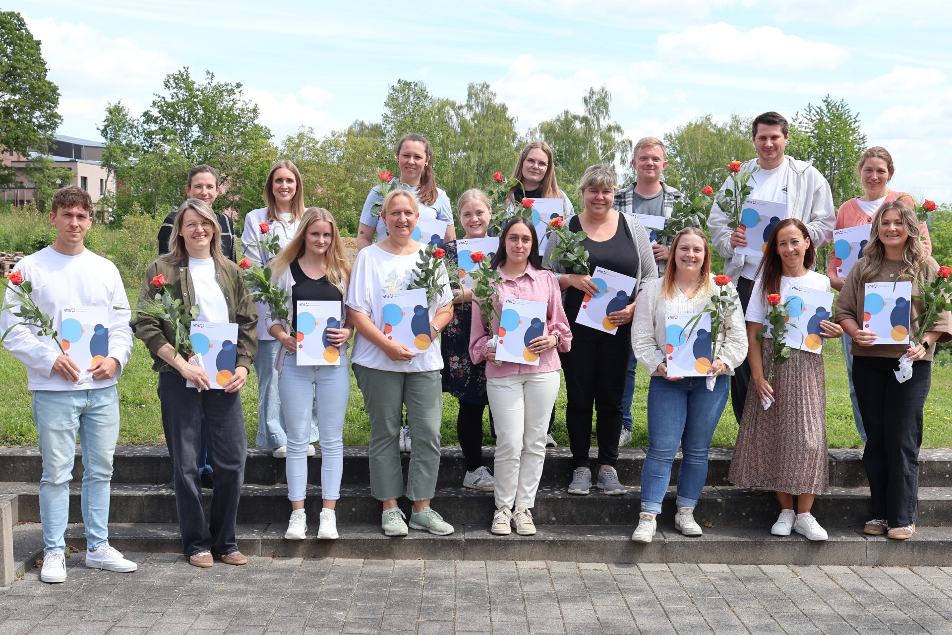 Eine Gruppe von Menschen steht mit Zertifikaten und Blume in der Hand auf einer Außentreppe.