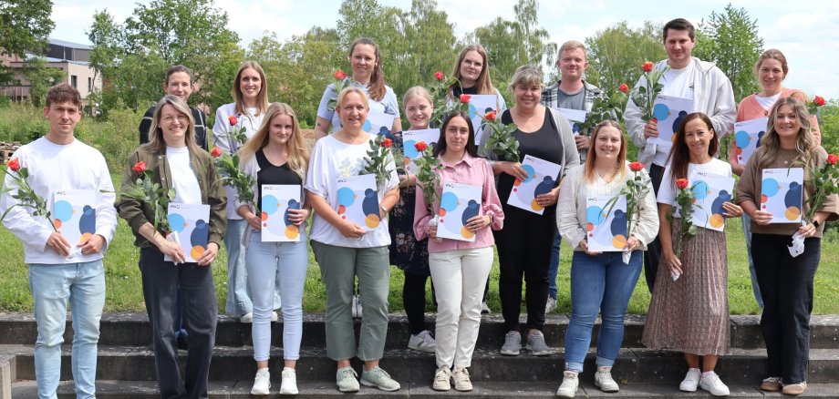 Eine Gruppe von Menschen steht mit Zertifikaten und Blume in der Hand auf einer Außentreppe.